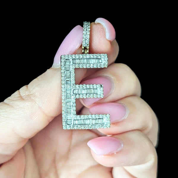 Diamond-encrusted letter 'E' pendant held by a hand with pink nail polish against a black background.